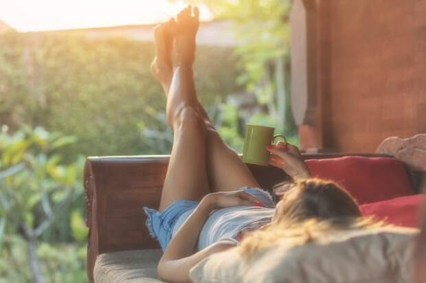 A woman lying down on a patio chair, looking at her sunny backyard, and holding a coffee mug.