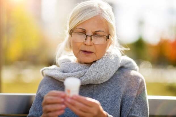 An older woman sitting on a sunny park bench and reading the label on a prescription pill bottle.