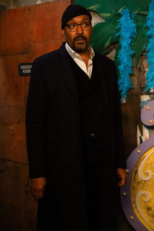 Alec standing by a dressing room door wearing a black trench coat and knit cap on The Irrational Season 2 Episode 3