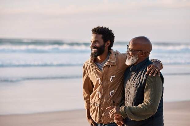 A man and his father smiling while standing on the beach with their arms around each other.