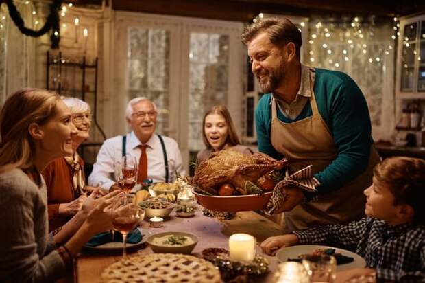 Group of smiling adults sitting around a Thanksgiving table full of food in a dining room decorated with twinkle lights while one person sets the cooked turkey down.