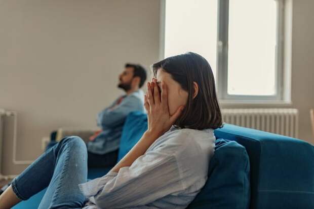A woman sitting at one end of a couch with her hands covering her face while a man sits with his arms crossed on the other end.