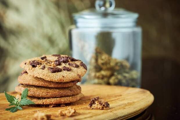 A stack of chocolate chip cookies sit on a wooden serving plate with a full glass cookie jar in the background.