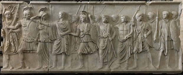 На барельефе когда-то был изображен император Домициан, потом его место занял император Нерва. Изображение преемника (четвертый слева) отличается от остальных фигур неправильным соотношением головы и тела