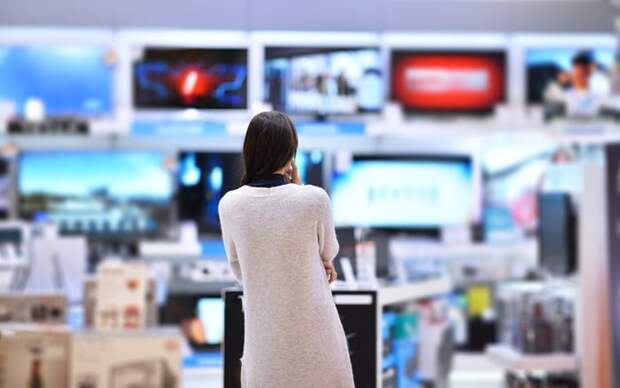 A woman standing in an electronics store facing a wall of TVs.