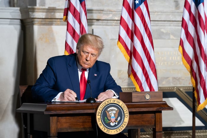 President Donald Trump signing a document while seated at a desk bearing the presidential seal.
