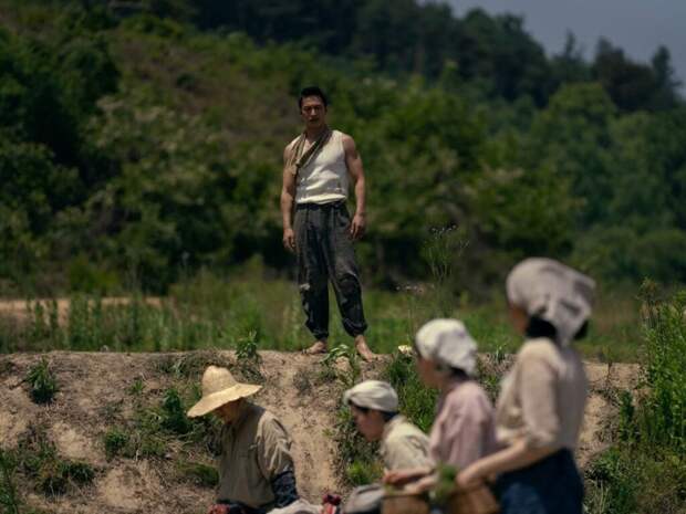 Mr. Kim in a sleeveless shirt stands on a bank over the working women