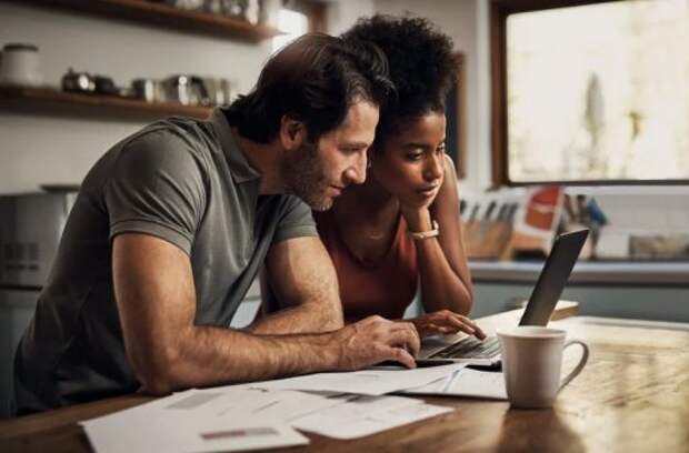 A man and woman sitting at their kitchen table with papers spread around them while researching something on a laptop.