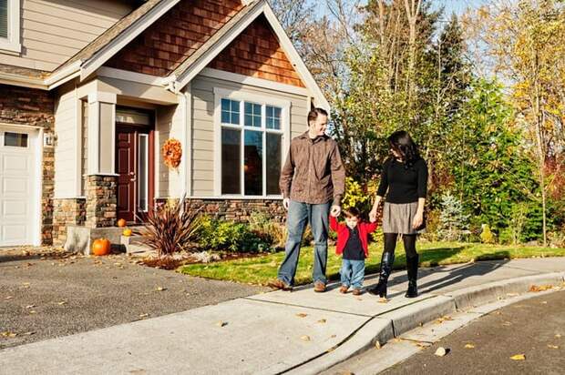 Two parents holding hands with their young child while walking down the sidewalk in front of a house.
