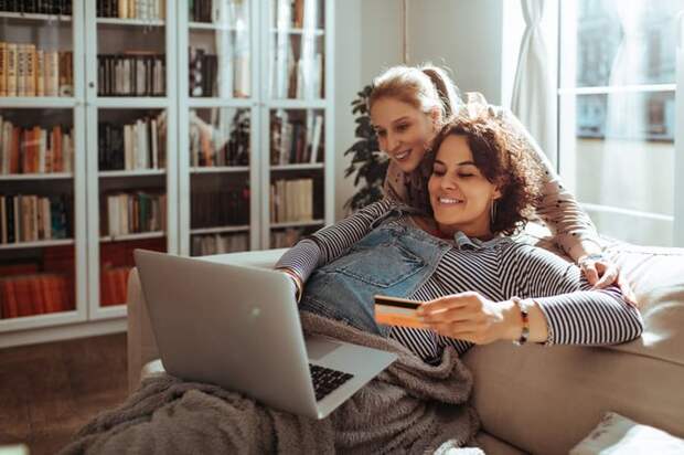 Pregnant woman holding credit card while using laptop with another woman looking over her shoulder.