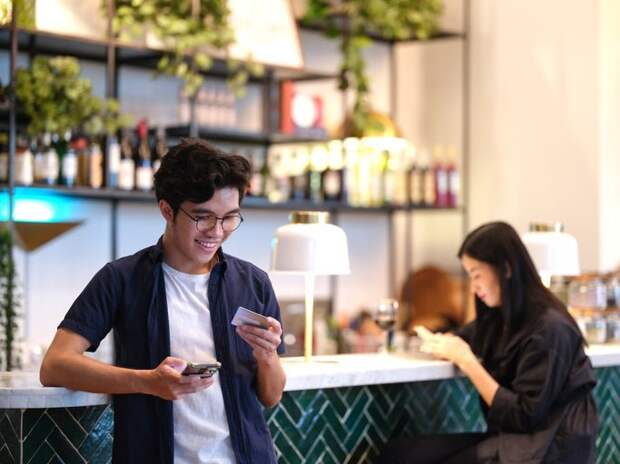 A man standing at a restaurant bar holding his phone and a credit card.