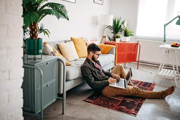 A person sitting on the floor leaning against a couch while typing on a laptop.