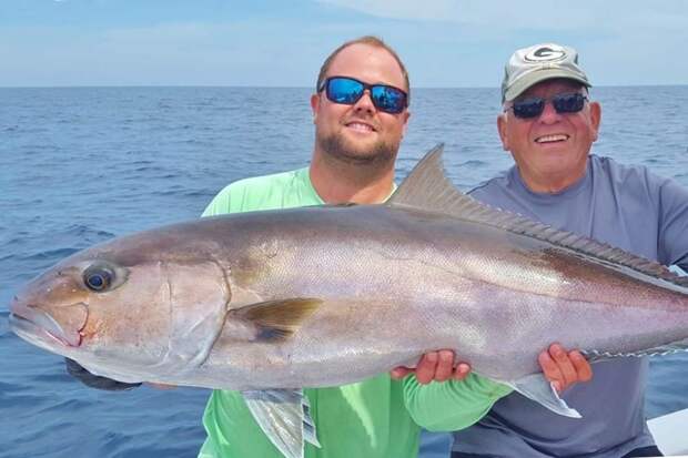 Two men hold a large Amberjack aboard a boat, with the ocean behind them
