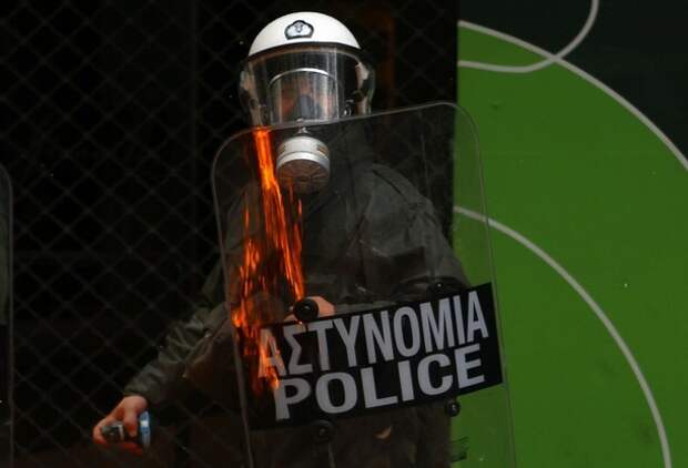 Fire is reflected on the shield of a policeman armed with a tear-gas canister during clashes in central Athens