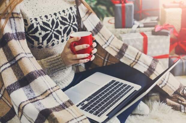 A woman holding a cup of coffee and a laptop with a blanket and presents around her.