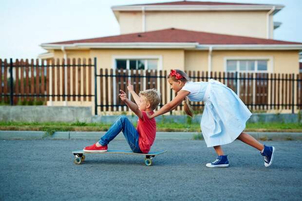 Happy girl pushing skateboard with her brother along fence of their new house