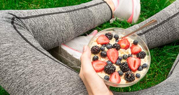 a hand holds a bowl of yogurt with fruit on someone's lap, the person is wearing running clothes, their torso is out of the frame