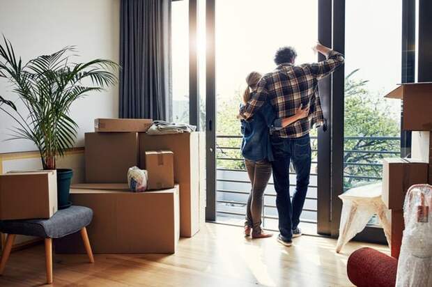 A man and woman standing with their arms around each other looking out a large window with moving boxes in the living room behind them.