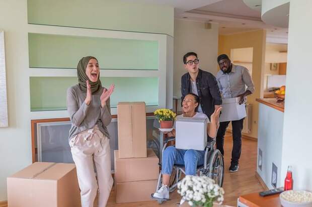Four adults, including one using a wheelchair, gasp in delight at their new home surrounded by moving boxes.
