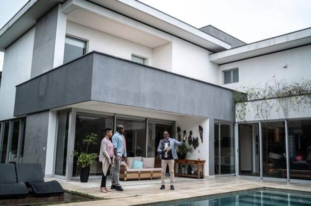 Two people speaking with a real estate agent while looking at a pool in the backyard of a house.