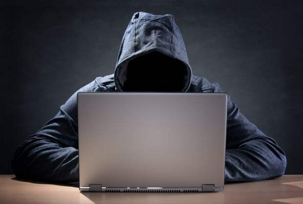 Hooded figure sitting at a laptop shrouded in darkness.