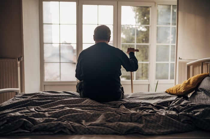 Person sitting on edge of bed, holding cane.