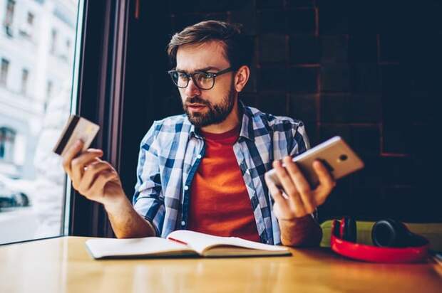 Man in blue checked shirt looks thoughtful with phone in one hand and credit card in the other.