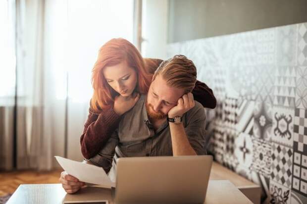 A woman leaning over a man's shoulder as he sits in front of a laptop with his head resting in his hand and holds up a piece of paper.