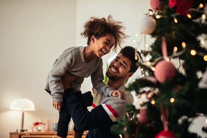 A smiling dad holding up his laughing daughter while they decorate a Christmas tree.