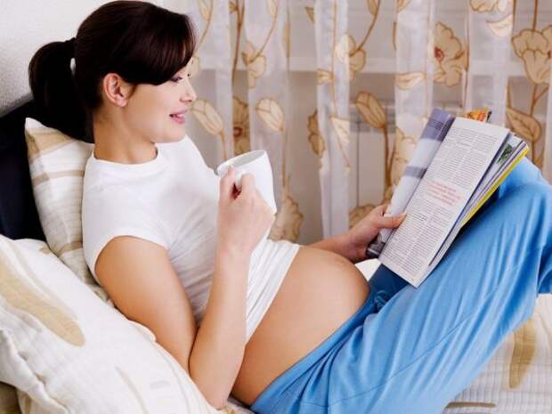 Беременная женщина в белой майке и голубых штанах с чашкой чая, облокотившись на подушки, читает журнал