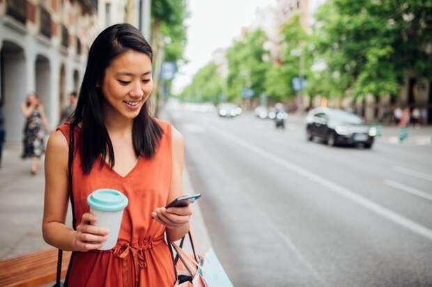 A young woman walking down the street with a coffee in one hand and looking at her phone in her other hand.