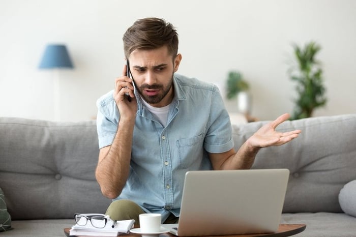 A man sitting on his couch and looking at his laptop in confusion while making a phone call.