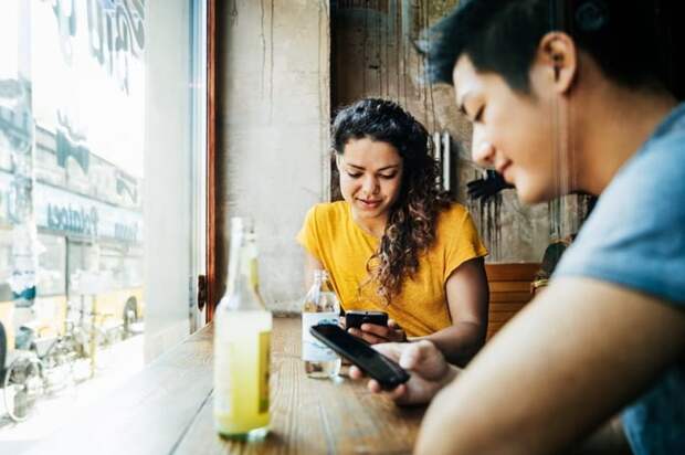 Two people sitting at a window table in a restaurant and looking at their cellphones.