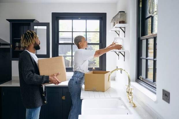 A couple unpacks cardboard boxes and organizes jars on shelves in their new kitchen.