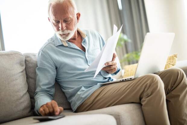 A person on a couch holding documents and using a calculator and laptop.