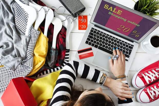 A woman shopping an online sale from her laptop while surrounded by clothes and shoes.