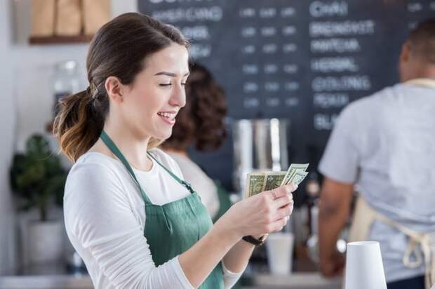 A smiling young barista counts cash in coffee shop.