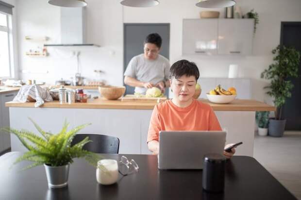 One spouse uses a laptop at the kitchen table while another prepares food in the background.